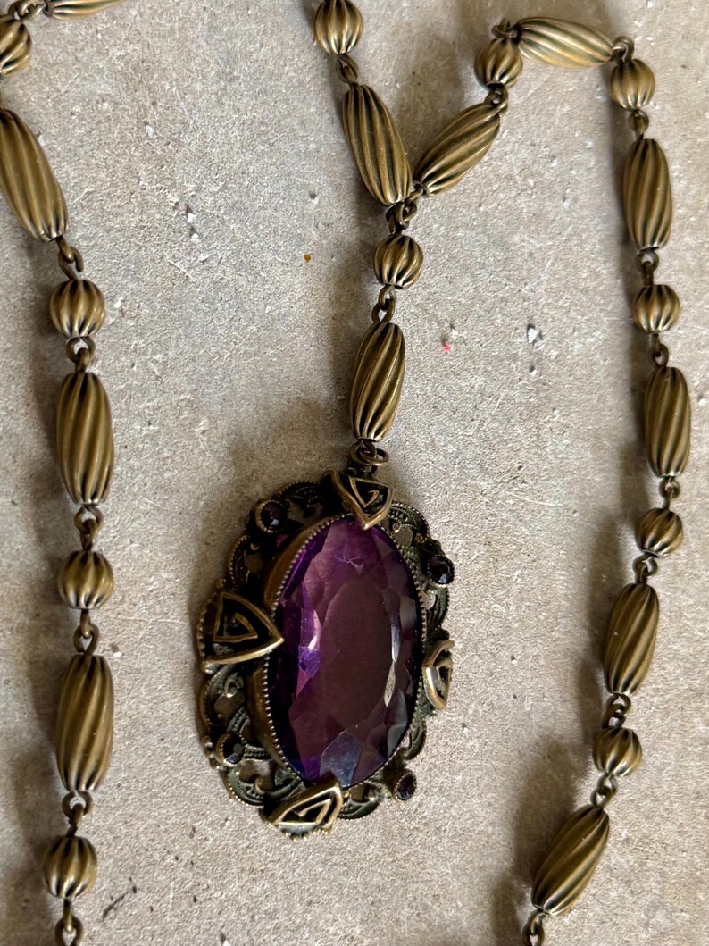 May include: Antique gold-tone necklace featuring an oval, faceted purple gemstone pendant. The pendant is set in an ornate filigree frame with geometric accents. The necklace chain has a series of ribbed and round beads.