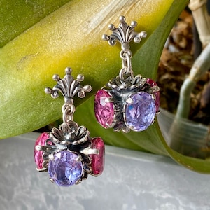 May include: A pair of silver dangle earrings with a floral design. Each earring features a large purple gemstone surrounded by three smaller pink gemstones.