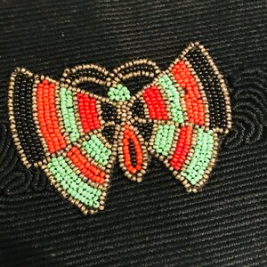 May include: A black fabric background with a beaded butterfly design. The butterfly is made of black, red, and green beads.