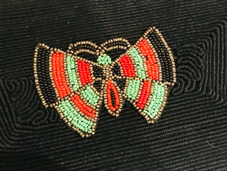 May include: A black fabric background with a beaded butterfly design. The butterfly is made of black, red, and green beads.
