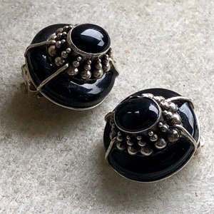 May include: A pair of black and silver clip-on earrings with a round, domed design. The earrings feature a black stone set in a silver bezel and surrounded by a ring of silver beads.