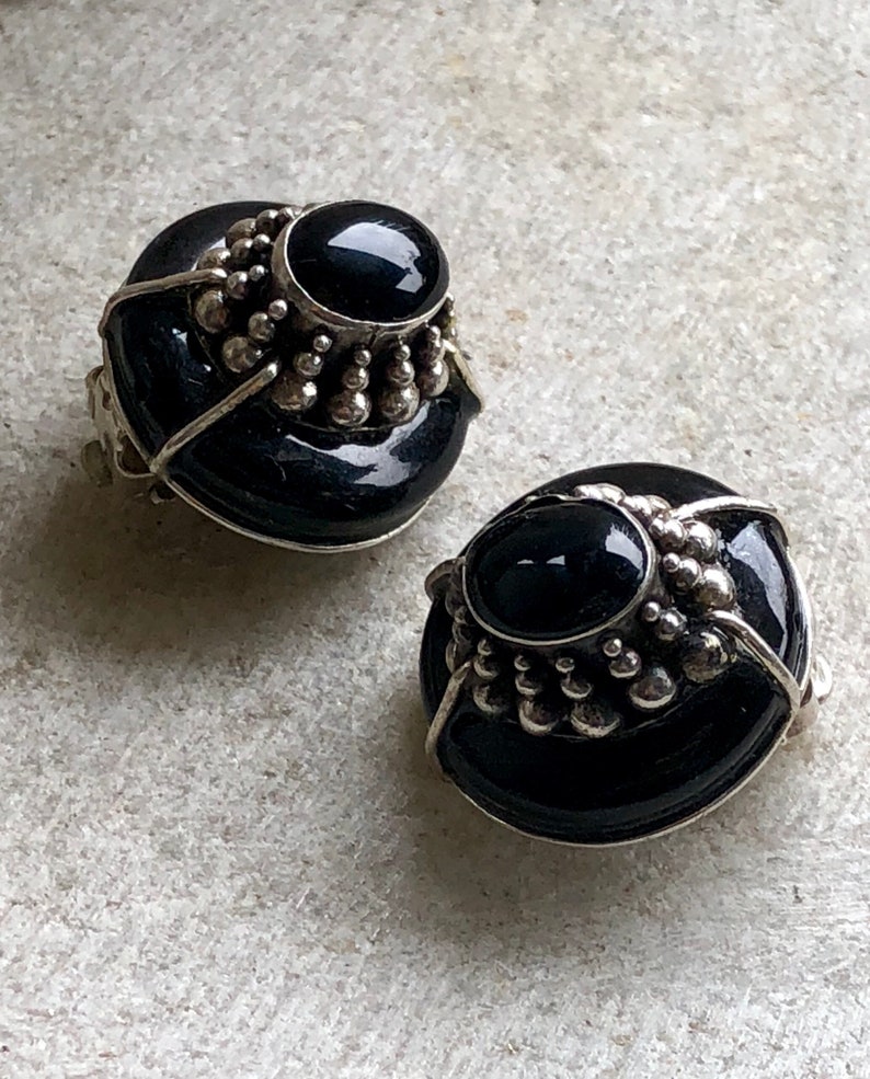 May include: A pair of black and silver clip-on earrings with a round, domed design. The earrings feature a black stone set in a silver bezel and surrounded by a ring of silver beads.