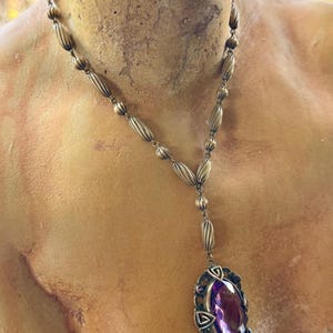 May include: Antique-style necklace with a bronze-colored chain and a large, oval, purple gemstone pendant. The pendant is framed by an ornate, filigree-style setting. The necklace is displayed on a bust.