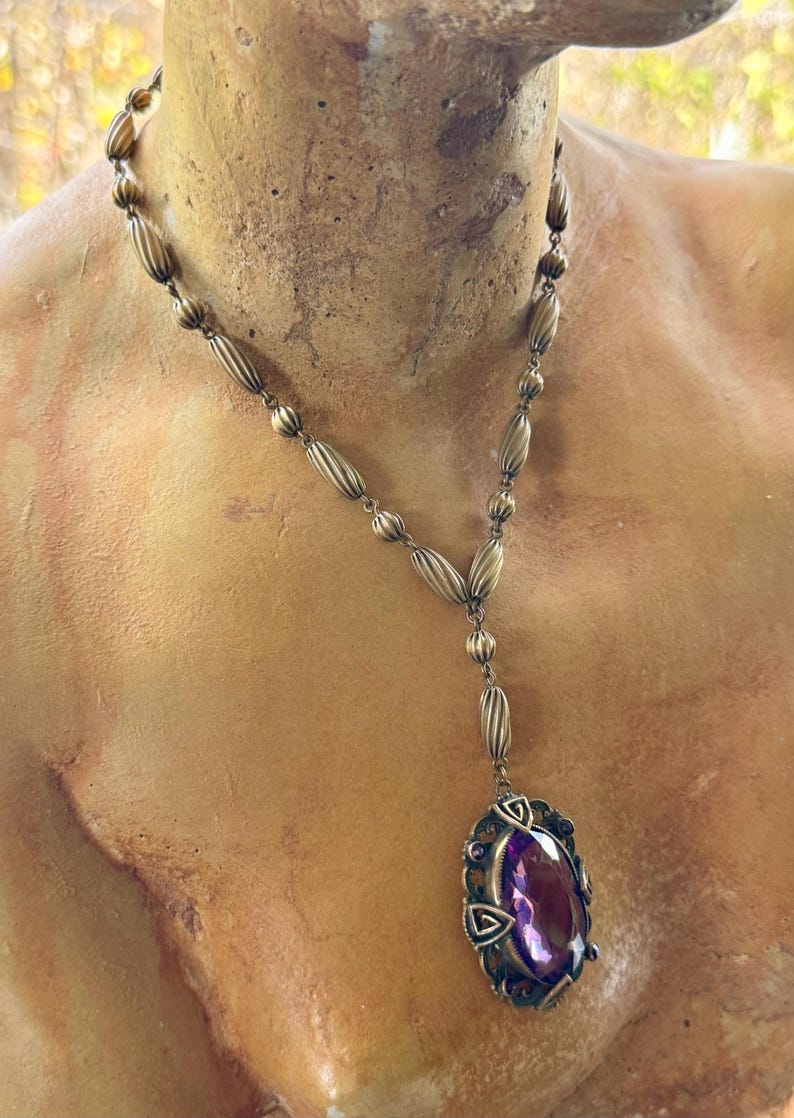 May include: Antique-style necklace with a bronze-colored chain and a large, oval, purple gemstone pendant. The pendant is framed by an ornate, filigree-style setting. The necklace is displayed on a bust.