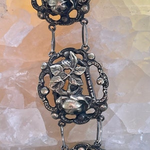 May include: A silver chain necklace with three decorative pendants featuring floral designs. Each pendant is intricately detailed with a rose and leaves.