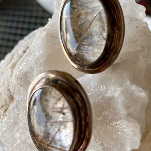 Puede incluir: Un par de pendientes de bot&oacute;n con un centro de cristal transparente y abovedado, que muestra delicadas inclusiones en forma de cabello. Los pendientes tienen un engaste de metal plateado con un borde redondeado. Se exhiben sobre una superficie cristalina blanca.