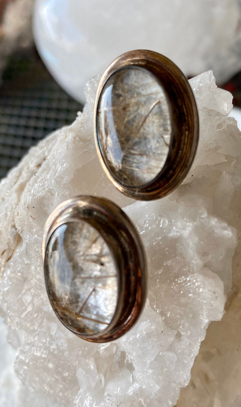 Puede incluir: Un par de pendientes de bot&oacute;n con un centro de cristal transparente y abovedado, que muestra delicadas inclusiones en forma de cabello. Los pendientes tienen un engaste de metal plateado con un borde redondeado. Se exhiben sobre una superficie cristalina blanca.