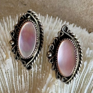 May include: A pair of silver earrings with pink mother of pearl oval insets. The earrings have a detailed, ornate design.