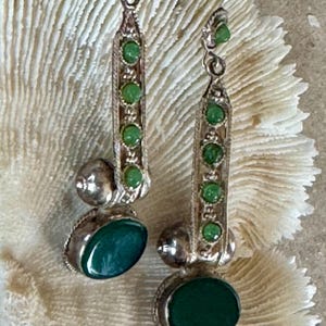 Pu&ograve; includere: Orecchini pendenti color argento con una serie di piccole pietre verdi e una pietra verde ovale pi&ugrave; grande nella parte inferiore. Gli orecchini hanno un design decorativo e ornato e sono sospesi a un gancio.