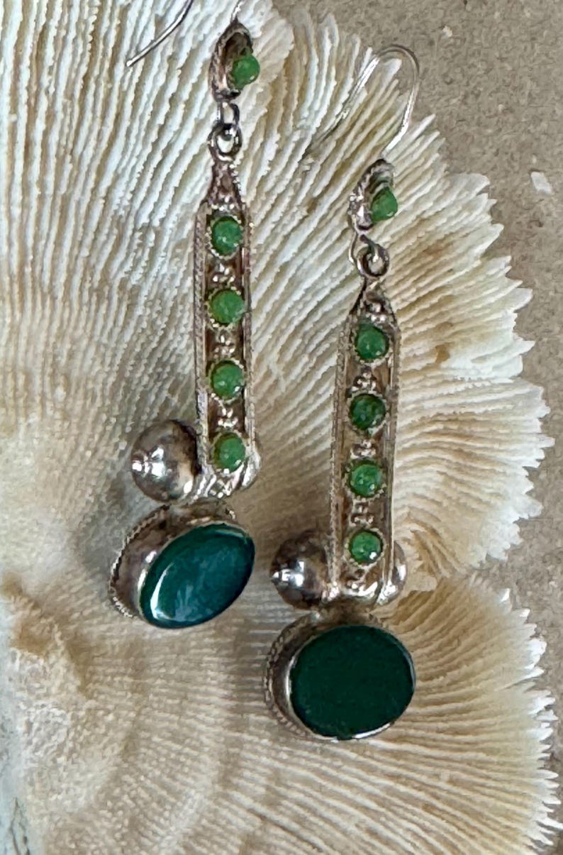 Pu&ograve; includere: Orecchini pendenti color argento con una serie di piccole pietre verdi e una pietra verde ovale pi&ugrave; grande nella parte inferiore. Gli orecchini hanno un design decorativo e ornato e sono sospesi a un gancio.