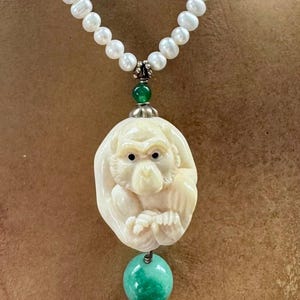 May include: A necklace featuring a strand of white, round, lustrous pearls. A carved, off-white monkey pendant is suspended from the pearls by a small green bead. A larger, smooth, round green bead hangs below the monkey carving as a focal point.