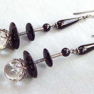 May include: A pair of silver earrings with black and clear glass beads. The earrings are made of silver wire and have a dangling design. The beads are round and have a smooth finish.