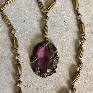 May include: Vintage brass-toned necklace featuring an oval pendant with a large, faceted purple gemstone. The pendant is framed by an ornate, filigree design with small purple accents. The necklace chain is composed of alternating ribbed and round beads.