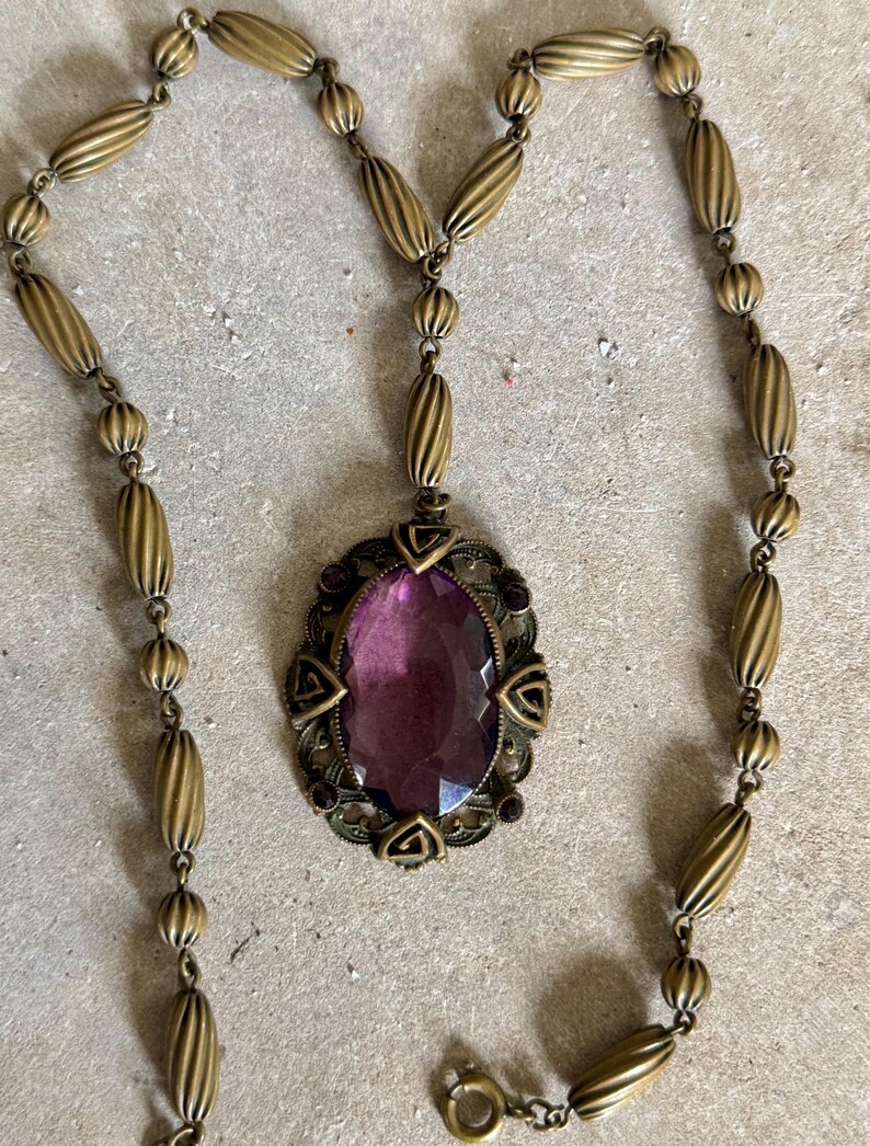 May include: Vintage brass-toned necklace featuring an oval pendant with a large, faceted purple gemstone. The pendant is framed by an ornate, filigree design with small purple accents. The necklace chain is composed of alternating ribbed and round beads.