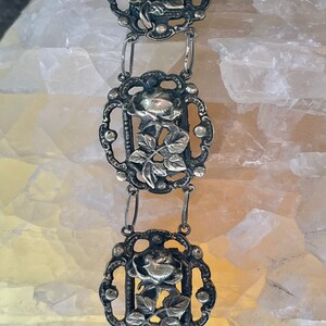 May include: A silver bracelet with a delicate design featuring three rose motifs. Each rose is surrounded by intricate filigree work and connected by small silver chains.