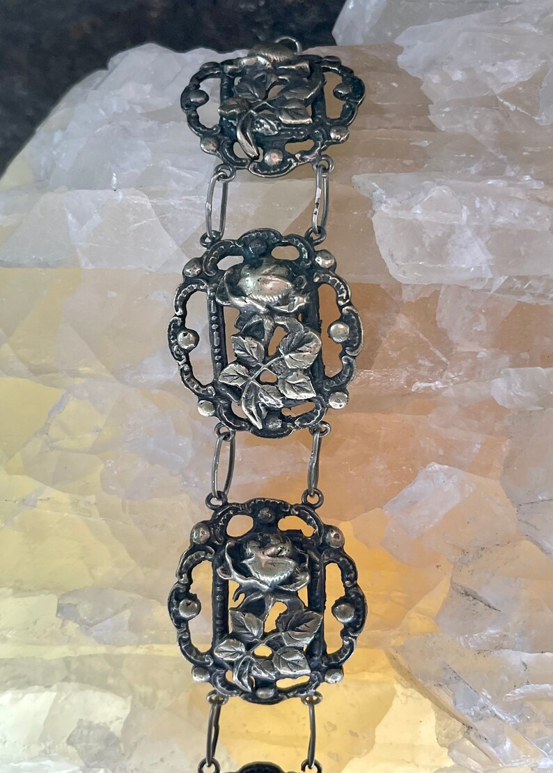 May include: A silver bracelet with a delicate design featuring three rose motifs. Each rose is surrounded by intricate filigree work and connected by small silver chains.