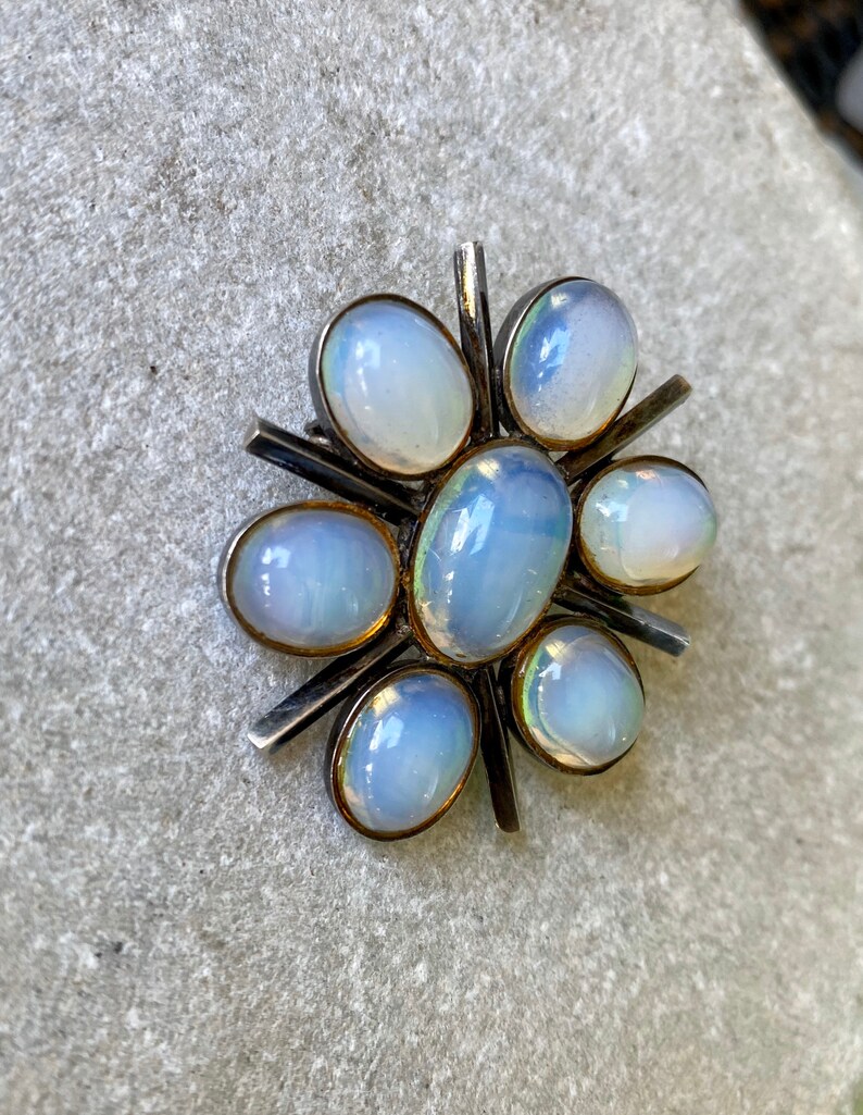 K&ouml;nnte beinhalten: Eine silberne Brosche mit einem Sternburst-Design mit sieben schillernden wei&szlig;en Opal-Steinen.
