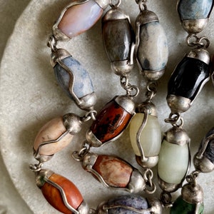 May include: A silver chain necklace with oval-shaped gemstone beads in various colors, including brown, red, blue, green, and white. The beads are set in silver.