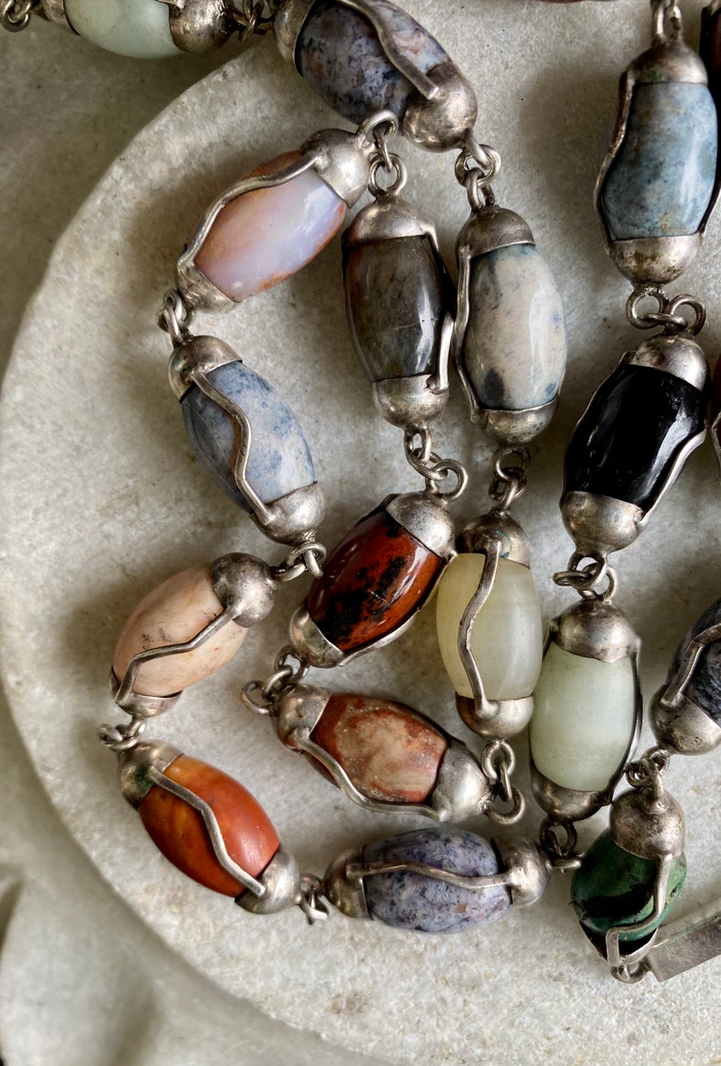 May include: A silver chain necklace with oval-shaped gemstone beads in various colors, including brown, red, blue, green, and white. The beads are set in silver.