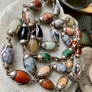 May include: A silver chain necklace with oval-shaped gemstone beads in various colors, including green, brown, red, and gray. The beads are set in silver.