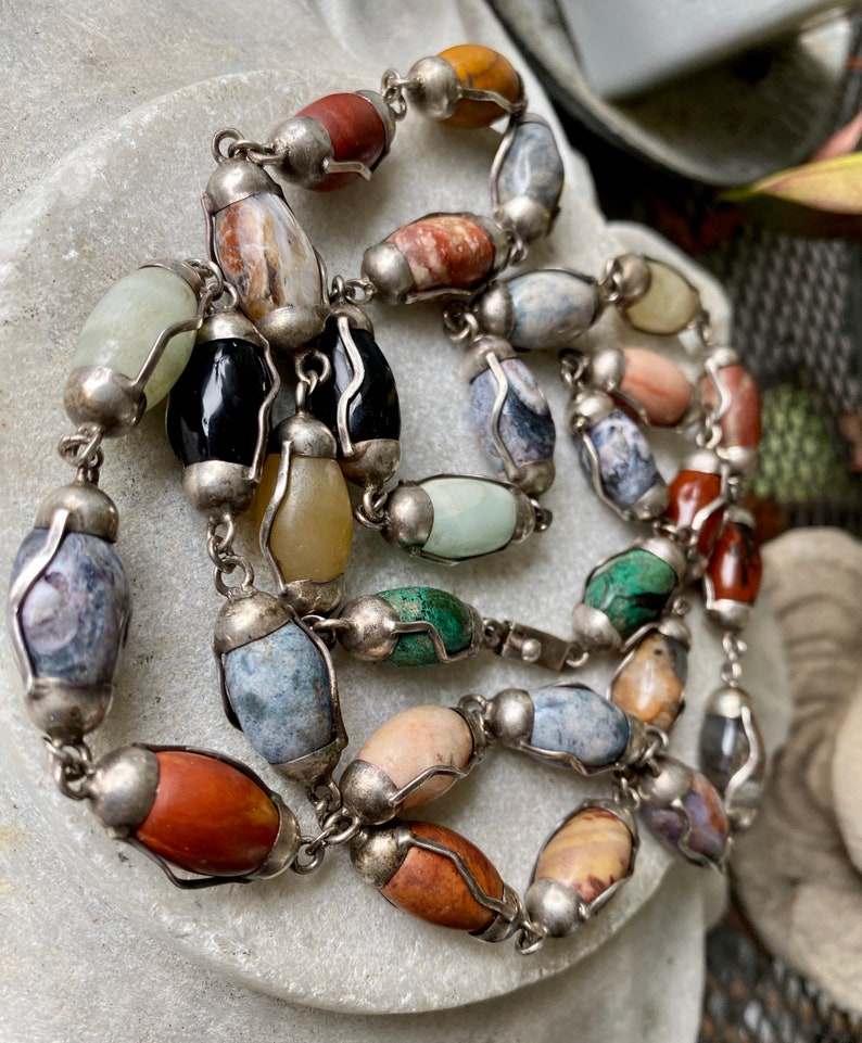 May include: A silver chain necklace with oval-shaped gemstone beads in various colors, including green, brown, red, and gray. The beads are set in silver.