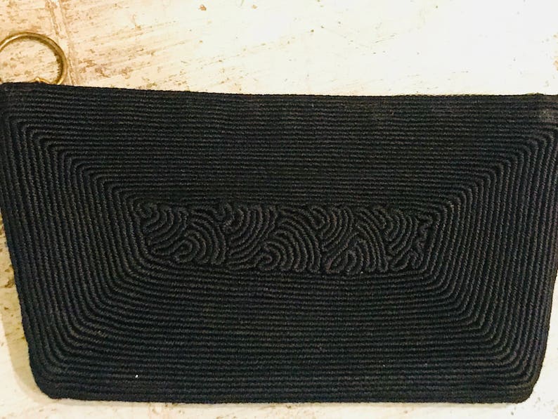 May include: A black woven clutch bag with a gold zipper pull. The bag has a textured design with a repeating pattern.