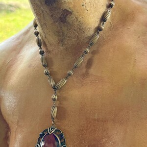 May include: Antique-style necklace with a large, oval, purple gemstone pendant. The pendant is framed by an ornate, filigree-style setting. The necklace chain features a series of elongated, ribbed beads. The metal has a vintage, tarnished appearance.