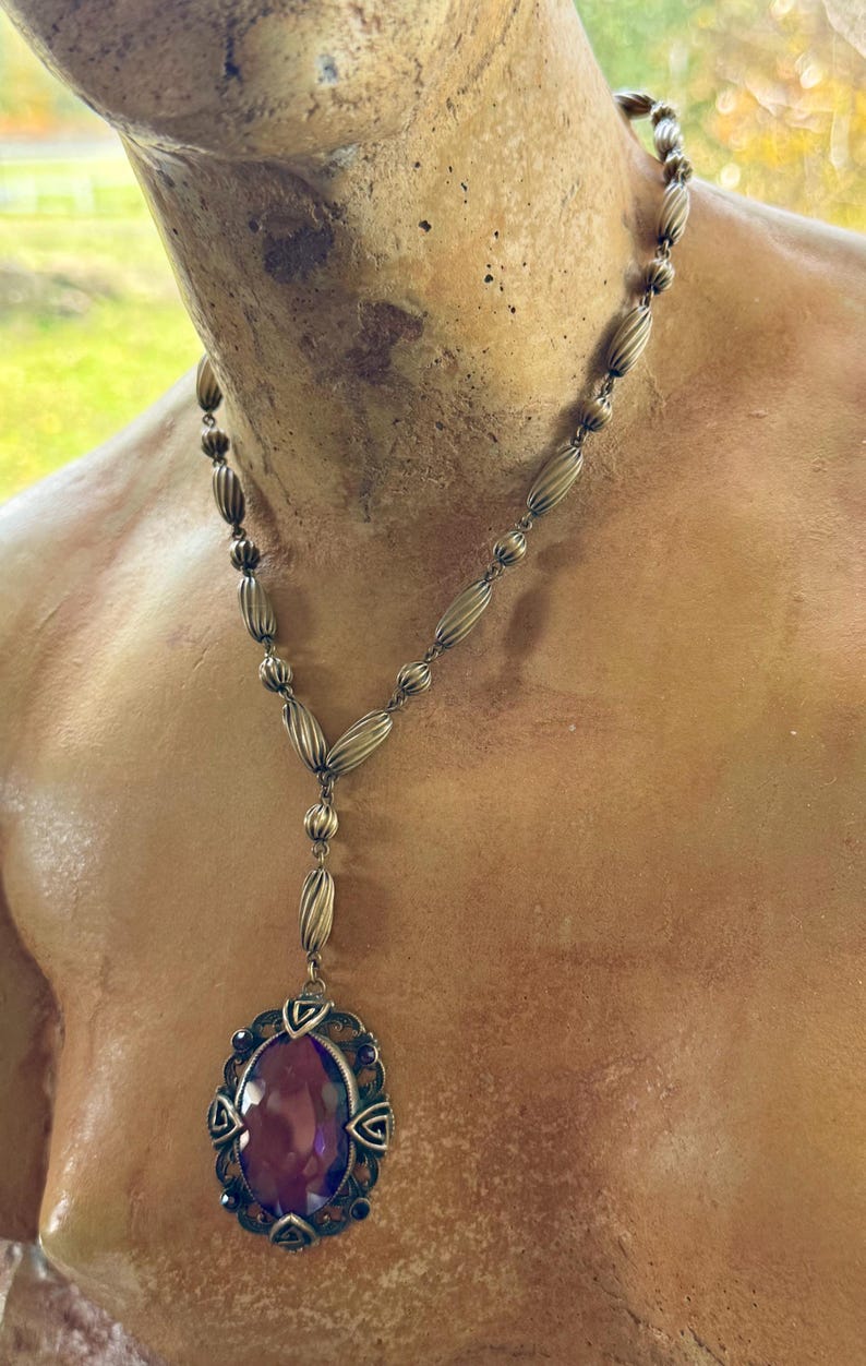 May include: Antique-style necklace with a large, oval, purple gemstone pendant. The pendant is framed by an ornate, filigree-style setting. The necklace chain features a series of elongated, ribbed beads. The metal has a vintage, tarnished appearance.