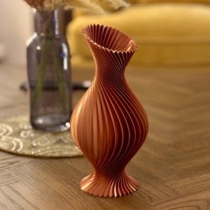 May include: A copper-colored ceramic vase with a unique, swirling, ribbed design. The vase is tall and slender with a wide opening at the top.