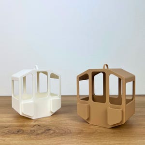 Pode incluir: Dois ornamentos decorativos geométricos em forma de cabine. Um é branco, o outro é castanho claro. Cada um tem uma forma retangular com janelas abertas e uma pequena argola no topo para pendurar. Os ornamentos têm um design moderno e minimalista.