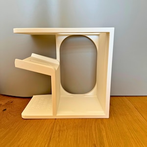 May include: White plastic shelf unit with a curved opening and a shelf on the side. The shelf unit is designed to hold items like a phone or a small plant.