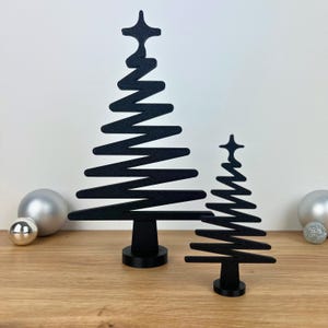 Può includere: Due decorazioni nere stilizzate a forma di albero di Natale. L'albero più grande ha un design a zig-zag e l'albero più piccolo ha un design simile. Entrambi gli alberi hanno una stella in cima e poggiano su una base rotonda. Ornamenti argentati sullo sfondo.
