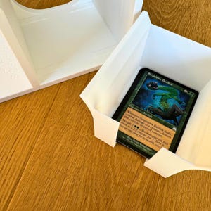May include: A white plastic card holder with a green and blue Magic: The Gathering card inside. The card is titled "Serpentine Basilisk" and has a green creature with a snake-like body on it.