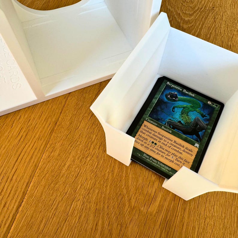 May include: A white plastic card holder with a green and blue Magic: The Gathering card inside. The card is titled "Serpentine Basilisk" and has a green creature with a snake-like body on it.