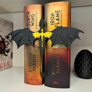 May include: A black dragon bookend with yellow wings holding up two books. The books are titled "Fourth Wing" and "Iron Flame" and are both by Rebecca Yarros. The books are on a shelf with a black dragon egg to the right.