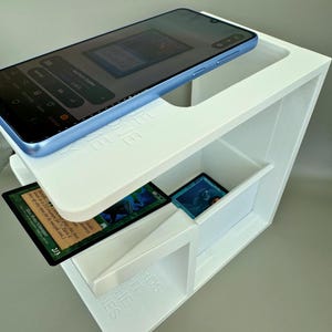 May include: White 3D printed phone and card holder with a blue phone on top and playing cards in the compartments. The holder has the words "THE NES" and "RDS" printed on the side.