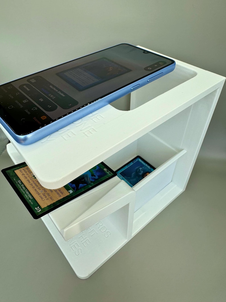 May include: White 3D printed phone and card holder with a blue phone on top and playing cards in the compartments. The holder has the words "THE NES" and "RDS" printed on the side.