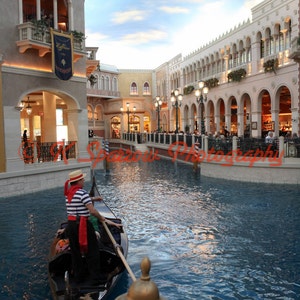 May include: A gondola glides through a canal, with a gondolier in a striped shirt and straw hat. Buildings with arches and balconies line the water's edge. The sky is blue with some clouds.