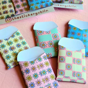 May include: A collection of small paper envelopes featuring colorful daisy patterns and checkered designs. Some envelopes display the text thestickergoblin. The envelopes are arranged in a group, showcasing various pastel color combinations including pink, blue, green, and yellow.