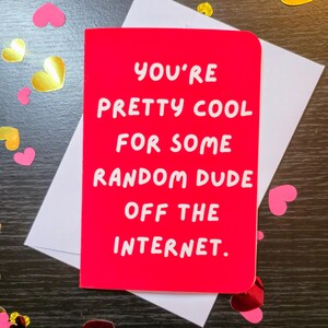 May include: A red greeting card with the text "YOU'RE PRETTY COOL FOR SOME RANDOM DUDE OFF THE INTERNET." sits on a white envelope. Pink and gold heart-shaped confetti is scattered around the card, creating a festive look.