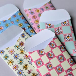 May include: Five small paper envelopes with white flaps, featuring various patterns including light blue, pink, and cream backgrounds with repeating daisy flower motifs and checkered designs in shades of pink, green, and yellow.