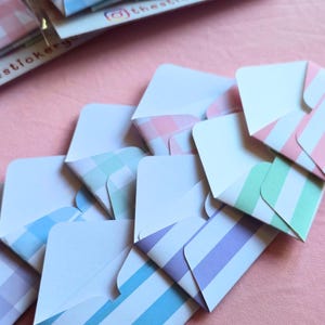 May include: A collection of small paper envelopes featuring various pastel-coloured patterns including blue, pink, green, and purple stripes and gingham checks. The envelopes are arranged in a scattered pile, with some packaged in clear plastic sleeves.