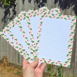 May include: A set of rectangular stationery sheets featuring a repeating pattern of green frogs and small red mushrooms on a light pink background. Each sheet has a white central area with horizontal blue lines for writing, arranged in a fan shape.