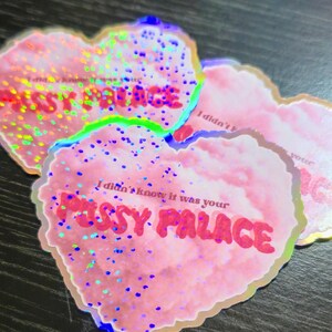 May include: Pink heart-shaped stickers with holographic glitter and the text "I didn't know it was your PUSSY PALACE" in red. The stickers have a glittery, iridescent effect and are arranged on a dark surface.