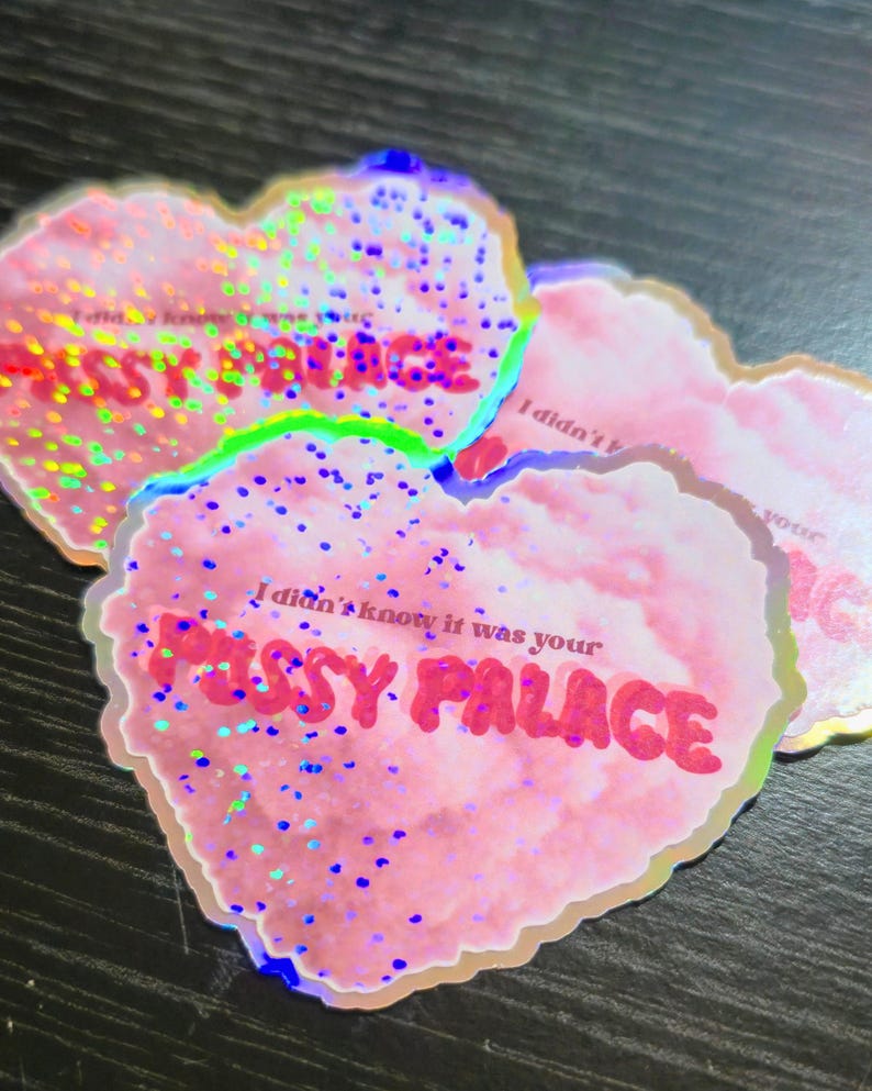 May include: Pink heart-shaped stickers with holographic glitter and the text "I didn't know it was your PUSSY PALACE" in red. The stickers have a glittery, iridescent effect and are arranged on a dark surface.