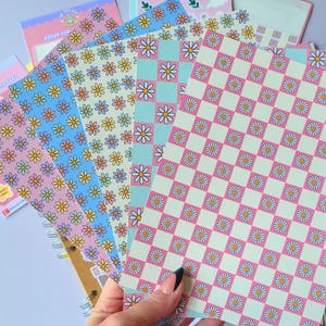 May include: A collection of rectangular sticker sheets featuring various patterns of small daisies. The designs include a pink and cream checkerboard pattern with daisies, a blue and pink checkerboard, and sheets with repeating daisy motifs.