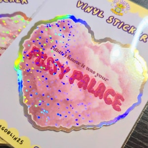 May include: A holographic vinyl sticker with a pink, cloud-like design and the text "FUSSY PALACE" in red. The sticker also includes the phrase "I didn't know it was your" in a smaller font. The sticker has a glittery effect and a scalloped edge.