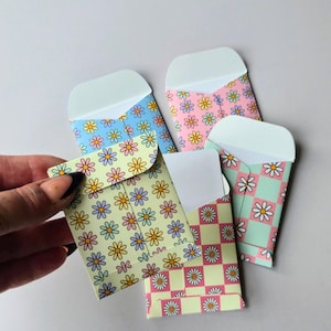 May include: A collection of small paper envelopes featuring various patterns including yellow, blue, and pink backgrounds with repeating daisy flower motifs and checkered designs in pastel colours. Each envelope has a rounded flap and white interior.
