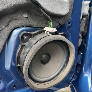 May include: A circular black car door speaker mounted within a blue metal panel. The speaker features a dark grey cone, a black centre dust cap, and a thick grey foam gasket around the perimeter. A white plastic electrical connector is attached.