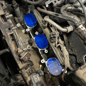 Può includere: Un blocco motore di un veicolo con tre coperture circolari blu per i condotti di aspirazione installate sulle aperture. I componenti metallici circostanti sono di colore grigio metallizzato e mostrano polvere e sporcizia.